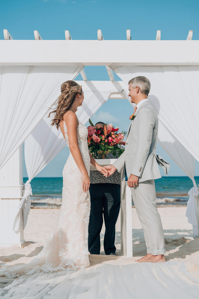 Bride and groom holding hands beneath a white beachfront wedding arbor at Secrets Maroma Beach Riviera Cancun.