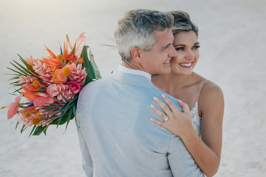 Bride and groom embrace on the beach at Secrets Maroma Beach Riviera Cancun, with the bride holding a vibrant tropical wedding bouquet.