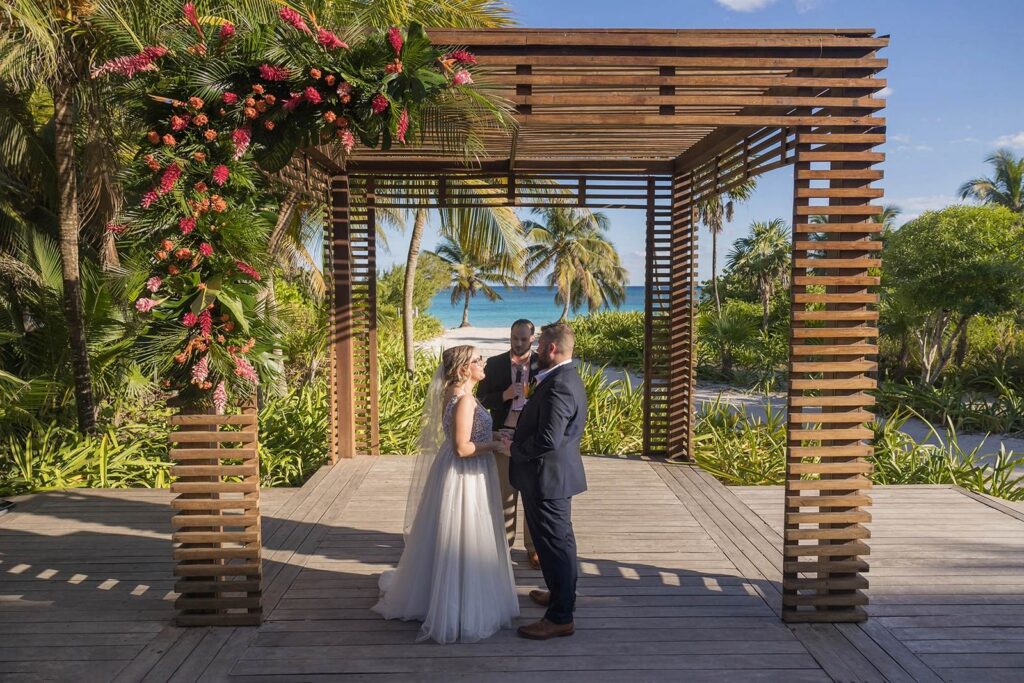 Bride and groom exchanging vows under a wooden gazebo with tropical florals at a destination wedding venue in Mexico