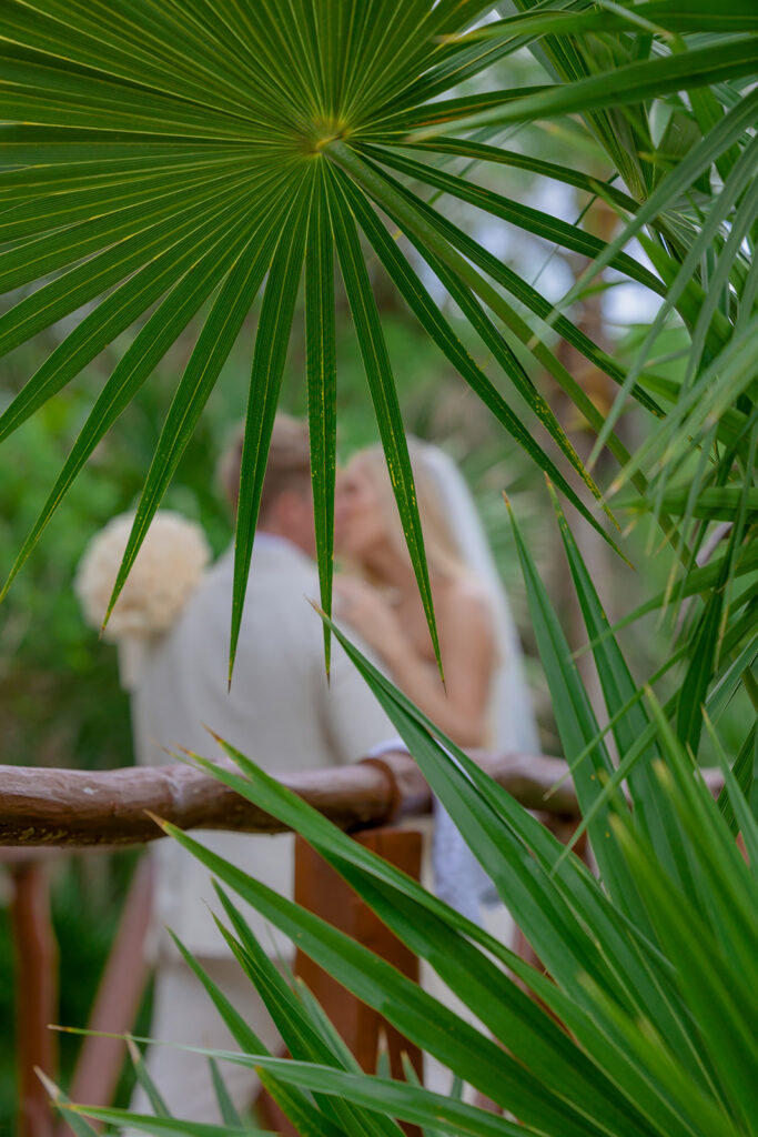 Bride and groom sharing a romantic moment framed by tropical palm leaves during a destination wedding in Mexico.