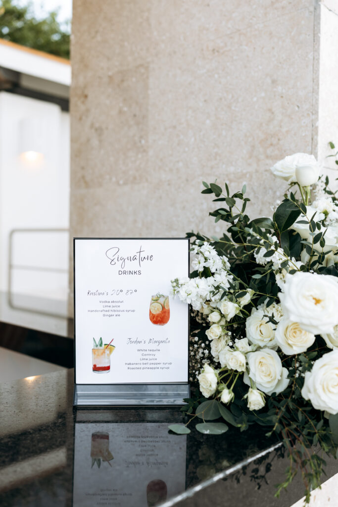 Signature wedding cocktail menu displayed with white floral arrangements at a destination wedding reception.