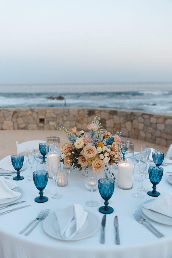 Oceanfront destination wedding reception table with candles, blue glassware, and floral centerpiece.